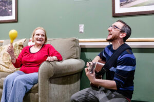 A music therapy session with a senior woman shaking a maraca beside Eric who's playing the guitar